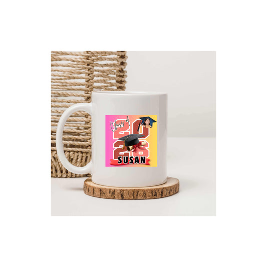 White decorative personalized mug featuring the graduate's name, custom photo, graduation year, and classic cap-and-diploma artwork with graduation design and name 'Susan' with a colorful design and text and the mug is sitting on a round wood-like coaster in front of a woven basket on a white background