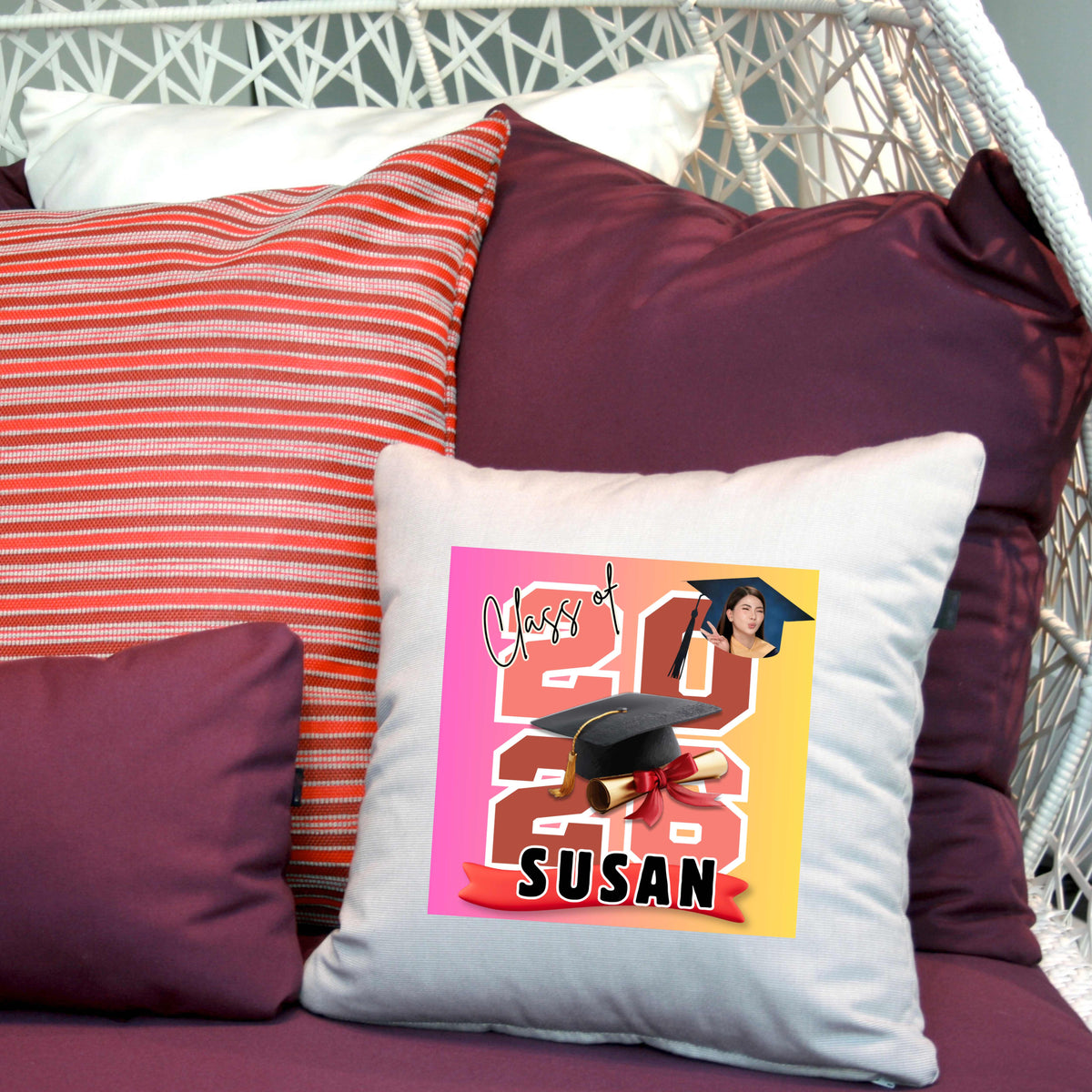 Decorative personalized pillow featuring the graduate's name, custom photo, graduation year, and classic cap-and-diploma artwork with graduation design and name 'Susan' on a chair with other pillows.