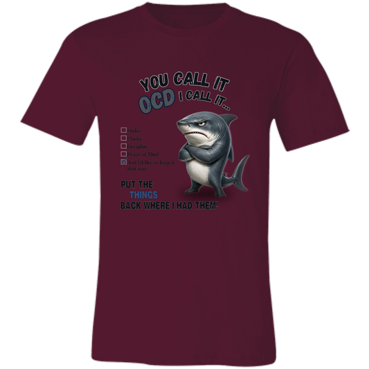 A unisex t-shirt in maroon, featuring a graphic print of a shark with crossed arms and the text 'YOU CALL IT OCD I CALL IT.' along with a checklist and the phrase 'PUT THE TRICKS BACK WHERE I HAD THEM!' on a transparent