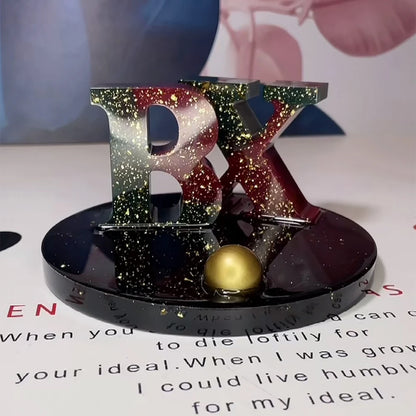 Decorative item with glittery 'BX' letters on a stand with a gold ball, placed on a surface with text.