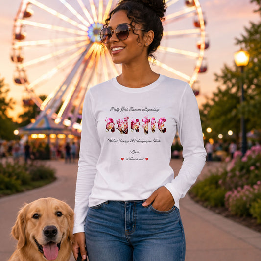 A woman wearing a white long-sleeve graphic t-shirt and blue jeans stands outdoors at a fairground with a Ferris wheel in the background, next to a golden retriever dog. The shirt features a graphic with the word 'AUNTIE' in pink floral letters and smaller black text above and below.