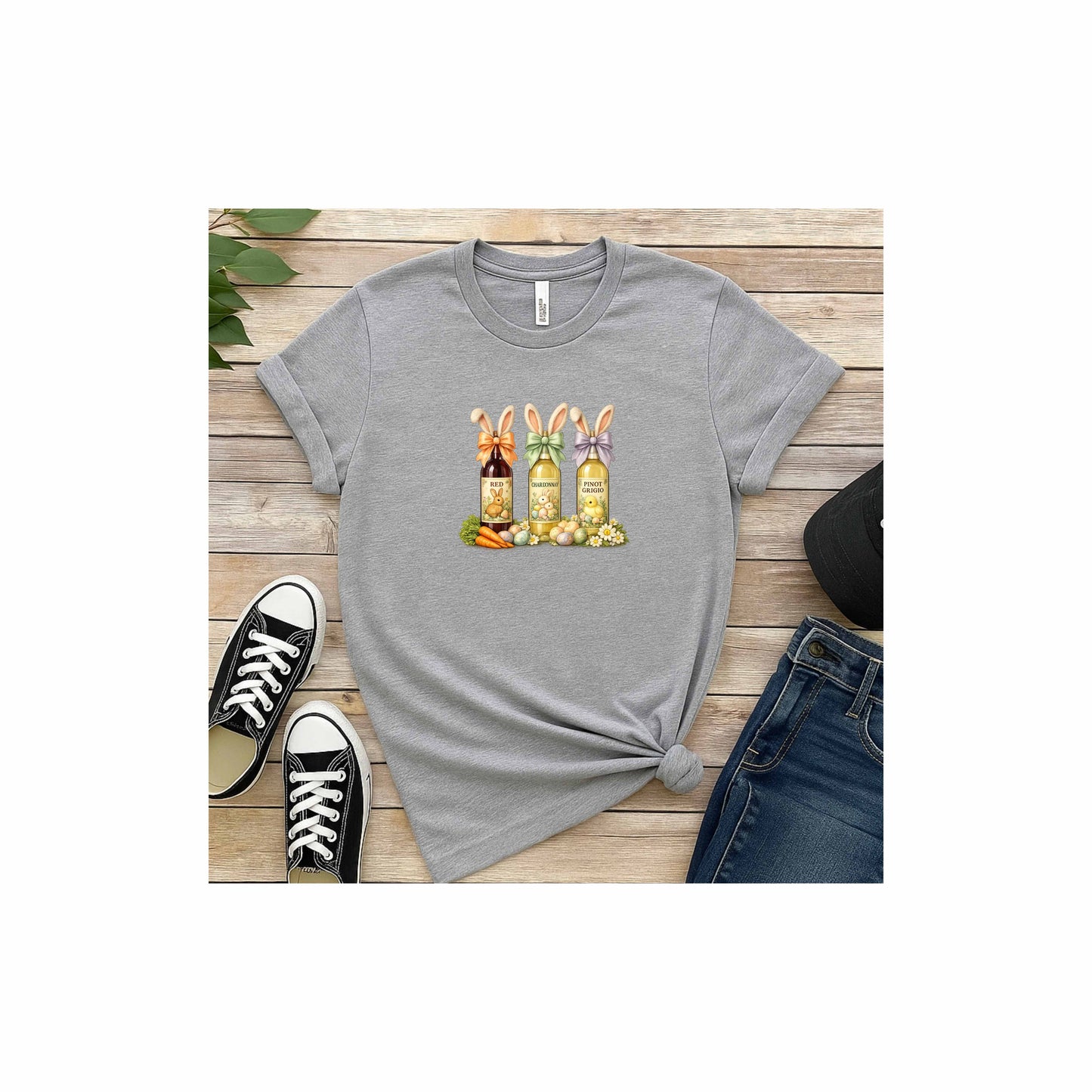 Mock-up image of a Gray t-shirt with a colorful graphic design of three wine bottles (Red, Chardonnay, Pinot Grigio) with bows around the neck of the bottle and bunny ears posed in different positions at the top, flowers, a bunch of carrots and easter designed eggs are at the bottom surrounding the bottoms.with a colorful graphic on a wooden surface with black and white shoes and jeans.