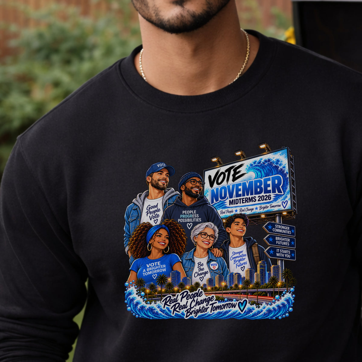 A close-up of a person wearing a black long-sleeve crewneck sweatshirt with a colorful political graphic on the front. The graphic features cartoon illustrations of five people wearing blue 'Vote November Midterms 2026' shirts, a city skyline, and directional signs.