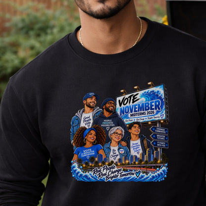 A close-up of a person wearing a black long-sleeve crewneck sweatshirt with a colorful political graphic on the front. The graphic features cartoon illustrations of five people wearing blue 'Vote November Midterms 2026' shirts, a city skyline, and directional signs.