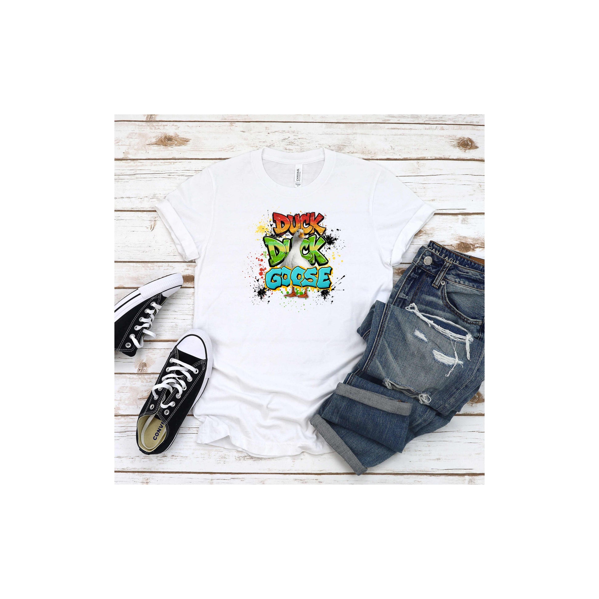 White t-shirt with 'Duck Duck Goose' graphic featuring a confident goose front and center surrounded by vibrant graffiti-inspired lettering and colorful paint splatter effects, black sneakers, and blue jeans on a wooden surface.