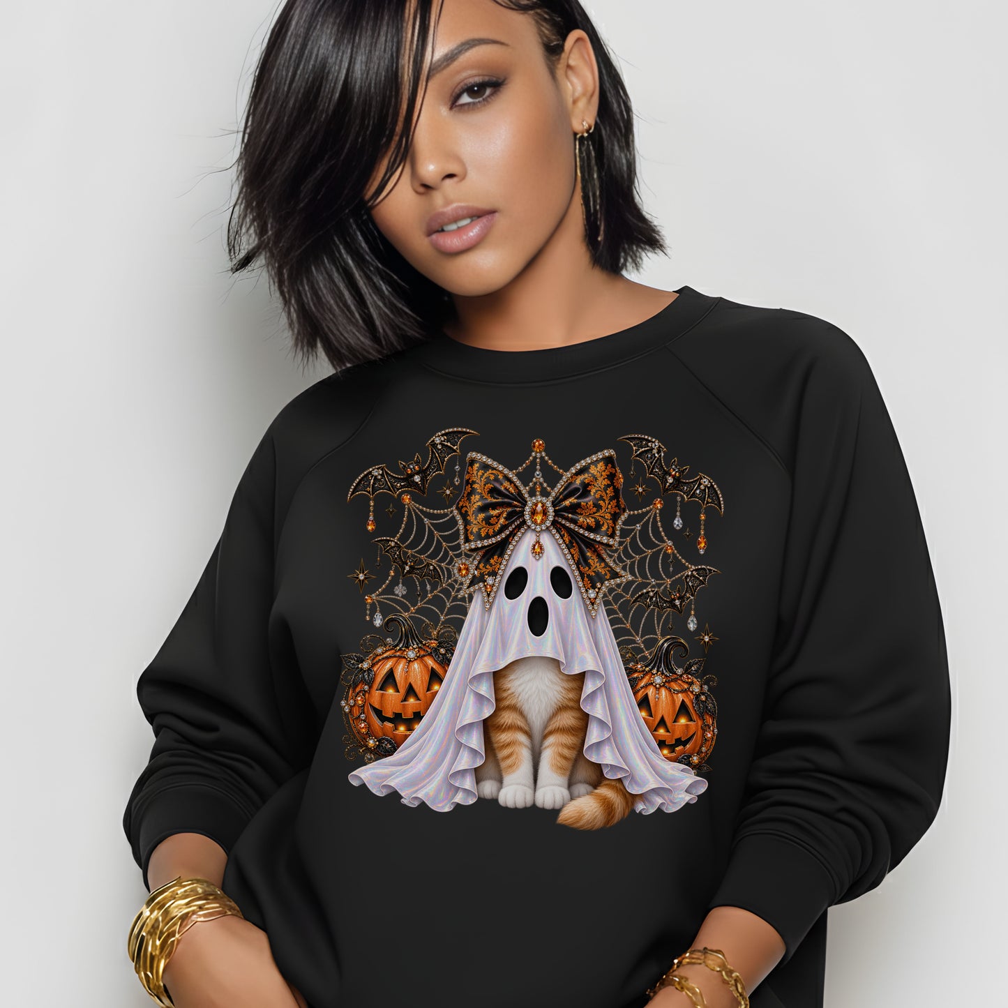 A woman wearing a black crewneck sweatshirt with a graphic of a ghostly cat surrounded by pumpkins, cobwebs, and a butterfly, set against a plain light gray background.