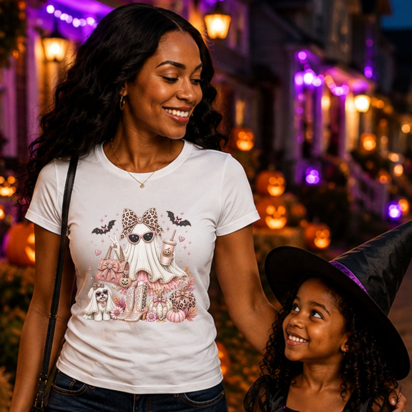 A woman and a child standing in a Halloween-lit street at night.  Women is wearing a white graphic T-shirt with a Halloween illustration of a Glamorous pearl-white ghost fashionista wearing sparkling sunglasses and a leopard-print bow with a dusty-rose handbag, embellished boots, rose-gold pumpkins, black bats, autumn leaves, and a small ghost dog companion.