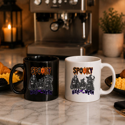 Two ceramic mugs with a Halloween graphic design, one black and one white, displayed on a marble countertop with a blurred coffee machine and candles in the background.
