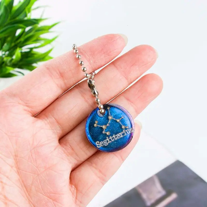 Hand holding a blue 'Sagittarius' zodiac pendant necklace with a plant in the background.