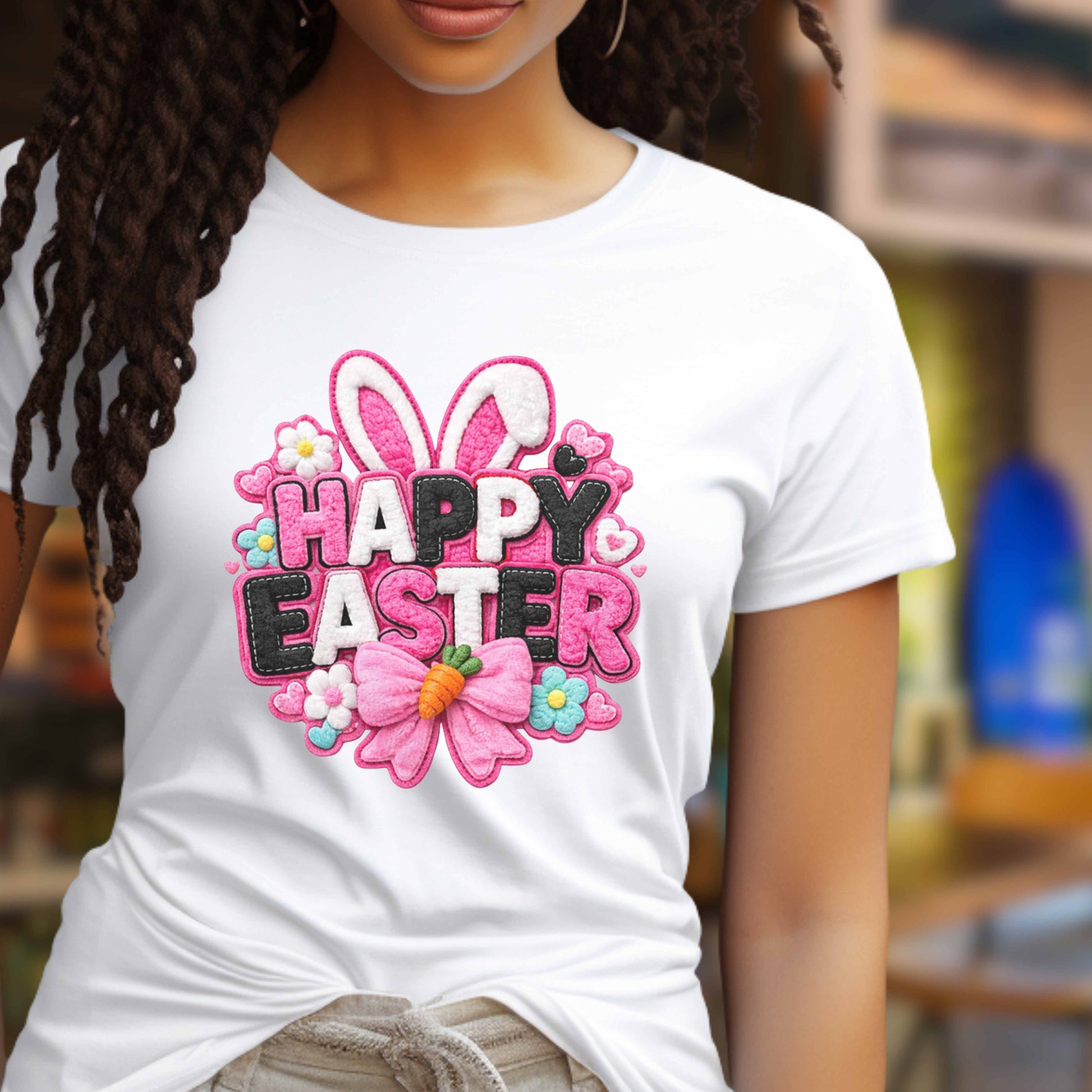 A woman wearing a white short-sleeve T-shirt with a colorful embroidered-style 'Happy Easter' graphic on the chest, featuring bunny ears, flowers, a carrot, and pink, white and black lettering, against a softly blurred indoor background.