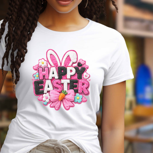 A woman wearing a white short-sleeve T-shirt with a colorful embroidered-style 'Happy Easter' graphic on the chest, featuring bunny ears, flowers, a carrot, and pink, white and black lettering, against a softly blurred indoor background.