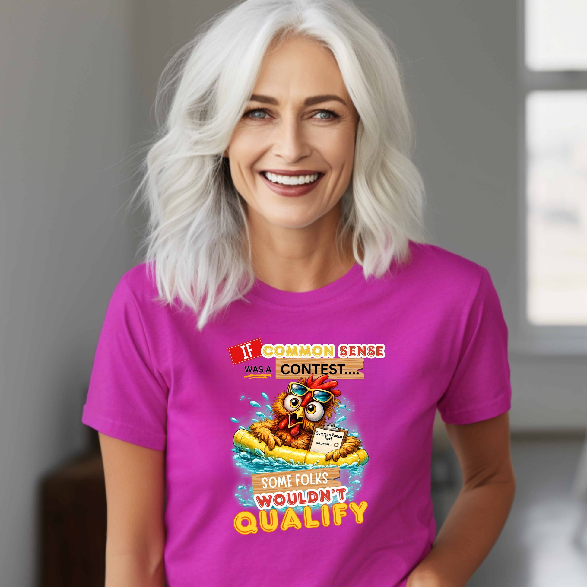 Woman wearing a purplish t-shirt with a colorful graphic and text featuring a wide-eyed pool-floating chicken desperately searching for common sense standing in a room with a window in the background