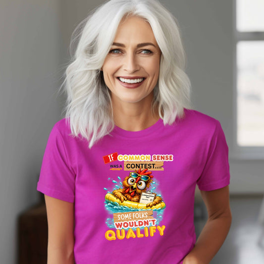 Woman wearing a purplish t-shirt with a colorful graphic and text featuring a wide-eyed pool-floating chicken desperately searching for common sense standing in a room with a window in the background