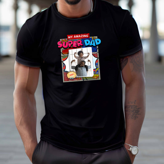 Person wearing a black t-shirt with 'My Amazing Super Dad' graphic design featuring vibrant superhero-inspired graphics, explosive comic panels, and a customizable photo area that has a father holding his son on his shoulders both showing their mussels on a transparent background