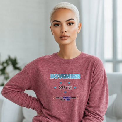 A woman with platinum blonde hair wearing a heathered dusty rose long-sleeve sweatshirt with a 'November Vote' graphic on the front, posed in a bright, modern room.