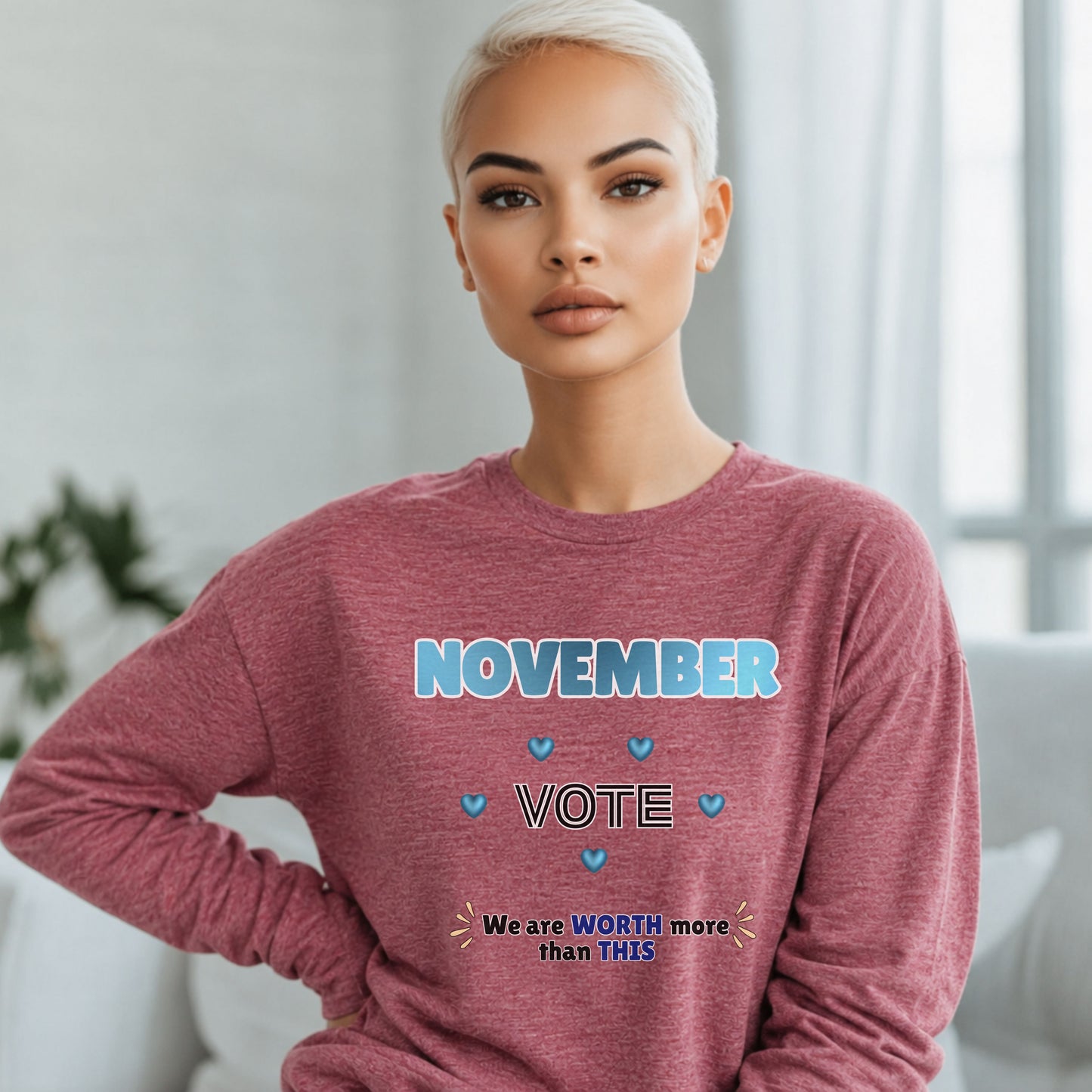 A woman with short platinum blonde hair wearing a heathered dusty rose long-sleeve sweatshirt with a blue and white graphic that reads 'NOVEMBER VOTE' and 'We are WORTH more than THIS'. She is posed indoors against a soft-focus light gray background.