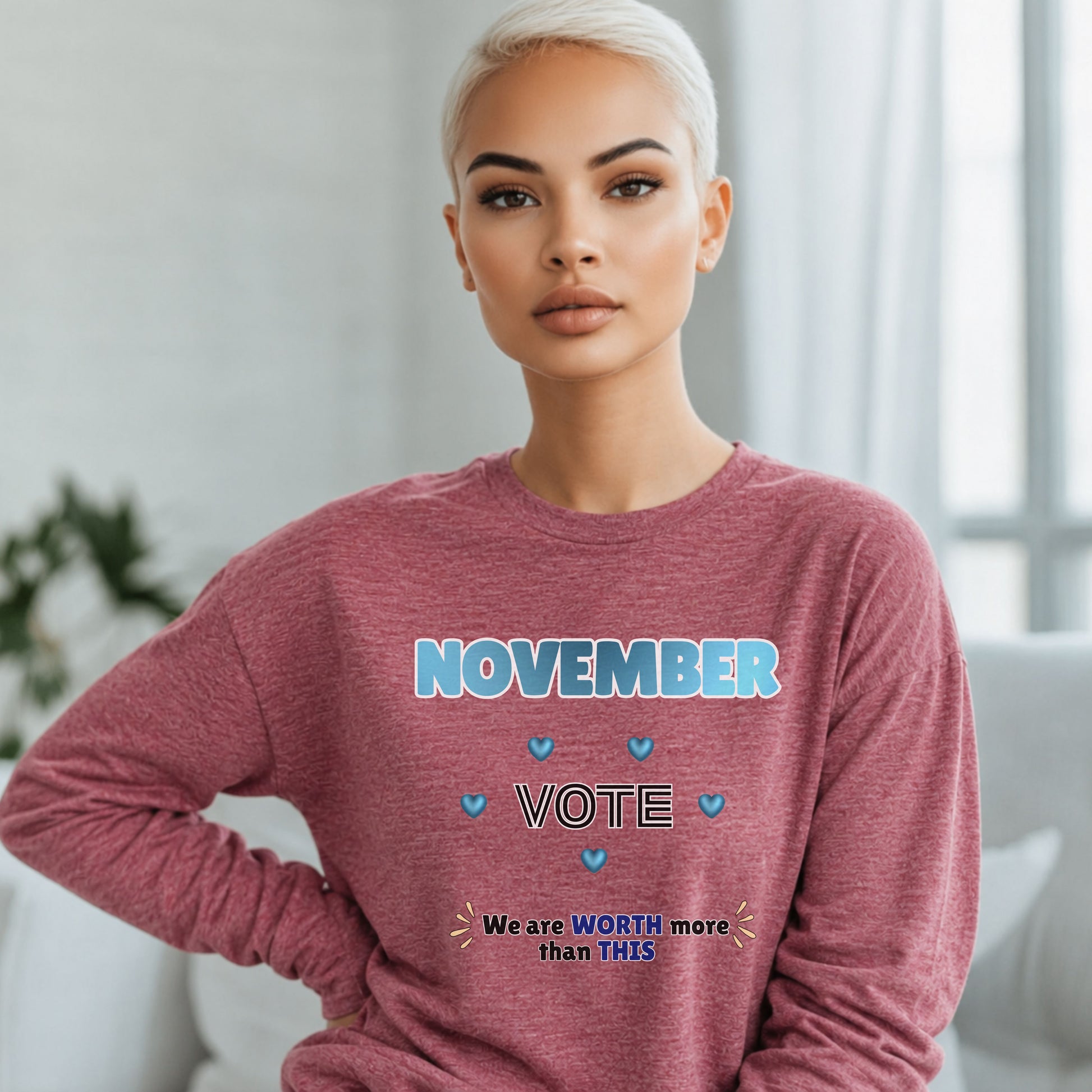 A woman with short platinum blonde hair wearing a heathered dusty rose long-sleeve sweatshirt with a blue and white graphic that reads 'NOVEMBER VOTE' and 'We are WORTH more than THIS'. She is posed indoors against a soft-focus light gray background.