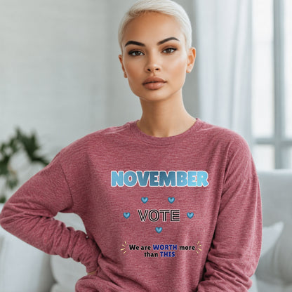 A woman with short platinum blonde hair wearing a heathered dusty rose long-sleeve sweatshirt with a blue and white graphic that reads 'NOVEMBER VOTE' and 'We are WORTH more than THIS'. She is posed indoors against a soft-focus light gray background.