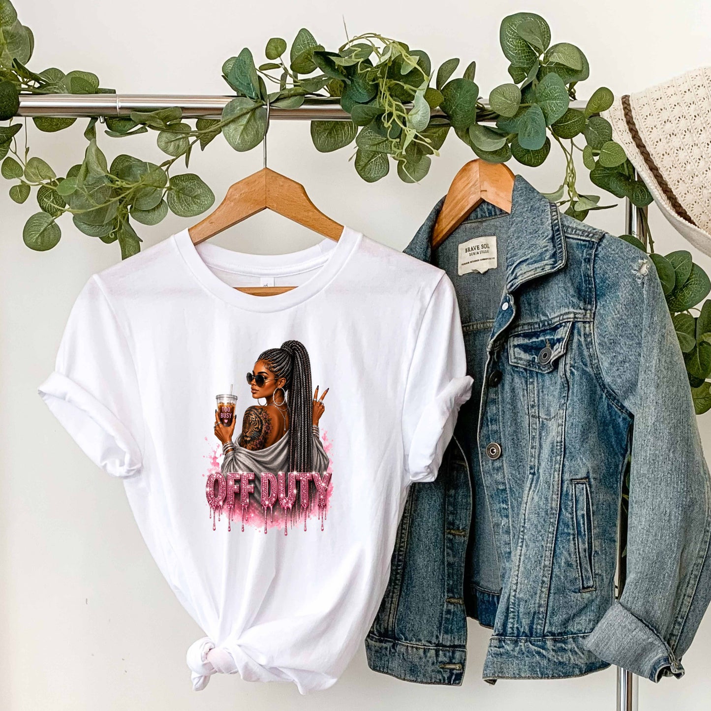 White t-shirt with a graphic design featuring a fashionable woman with a sleek braided ponytail accented with hints of silver, oversized sunglasses, statement jewelry, a bold tiger tattoo, and her favorite iced coffee in hand that says 'TOO BUSY' with a pink heart and sparkling pink rhinestone-inspired 'OFF DUTY' lettering on a transparent background, paired with a denim jacket on a hanger against a white wall with greenery.