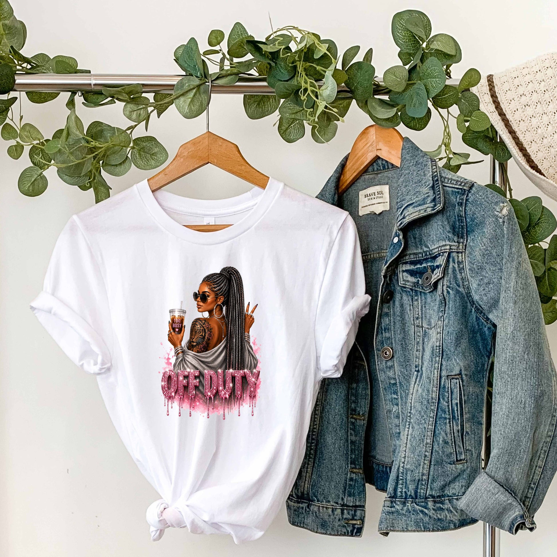 White t-shirt with a graphic design featuring a fashionable woman with a sleek braided ponytail accented with hints of silver, oversized sunglasses, statement jewelry, a bold tiger tattoo, and her favorite iced coffee in hand that says 'TOO BUSY' with a pink heart and sparkling pink rhinestone-inspired 'OFF DUTY' lettering on a transparent background, paired with a denim jacket on a hanger against a white wall with greenery.