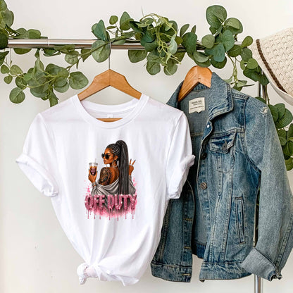White t-shirt with a graphic design featuring a fashionable woman with a sleek braided ponytail accented with hints of silver, oversized sunglasses, statement jewelry, a bold tiger tattoo, and her favorite iced coffee in hand that says 'TOO BUSY' with a pink heart and sparkling pink rhinestone-inspired 'OFF DUTY' lettering on a transparent background, paired with a denim jacket on a hanger against a white wall with greenery.