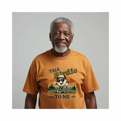 AI Generated African American older man wearing a mustard yellow t-shirt with a graphic design and text "Talk Birdie To Me" t-shirt featuring a cool cartoon golf ball rocking sunglasses on the green  leaning on a golf club and next to a putting hole with a flag inside of the hole with  a retro sunset background