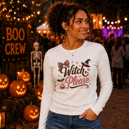 A woman wearing a white long-sleeve graphic t-shirt with 'Witch Please' written in a glittery pink and black font, featuring a witch hat and bow design. She is standing outdoors at a Halloween festival with pumpkins, a skeleton, and string lights in the background.