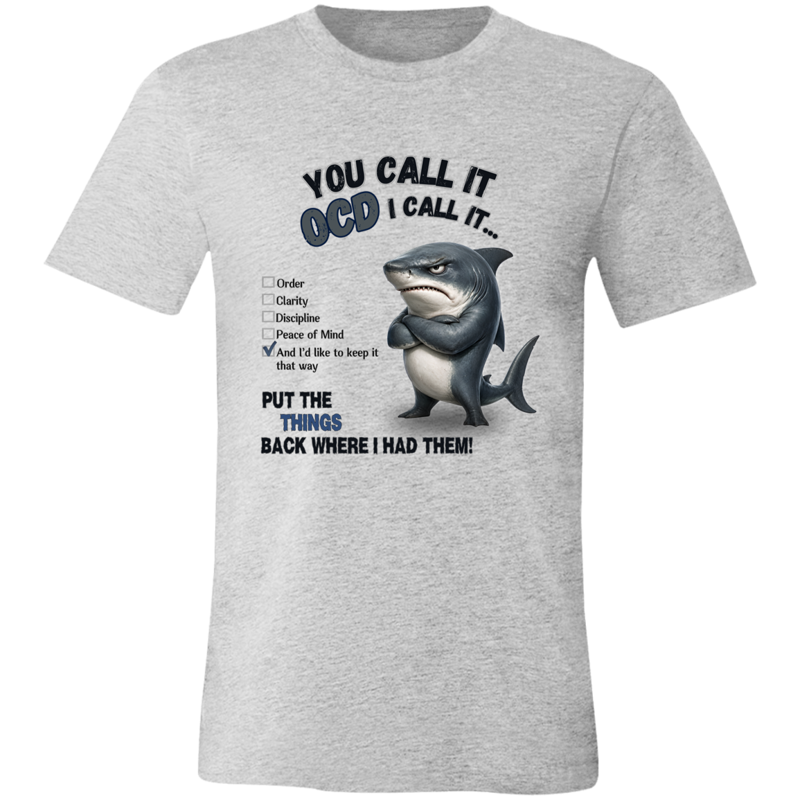 A unisex t-shirt in athletic heather, featuring a graphic print of a shark with crossed arms and the text 'YOU CALL IT OCD I CALL IT.' along with a checklist and the phrase 'PUT THE TRICKS BACK WHERE I HAD THEM!' on a transparent 