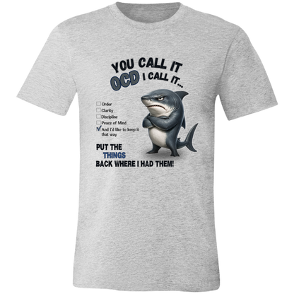 A unisex t-shirt in athletic heather, featuring a graphic print of a shark with crossed arms and the text 'YOU CALL IT OCD I CALL IT.' along with a checklist and the phrase 'PUT THE TRICKS BACK WHERE I HAD THEM!' on a transparent 