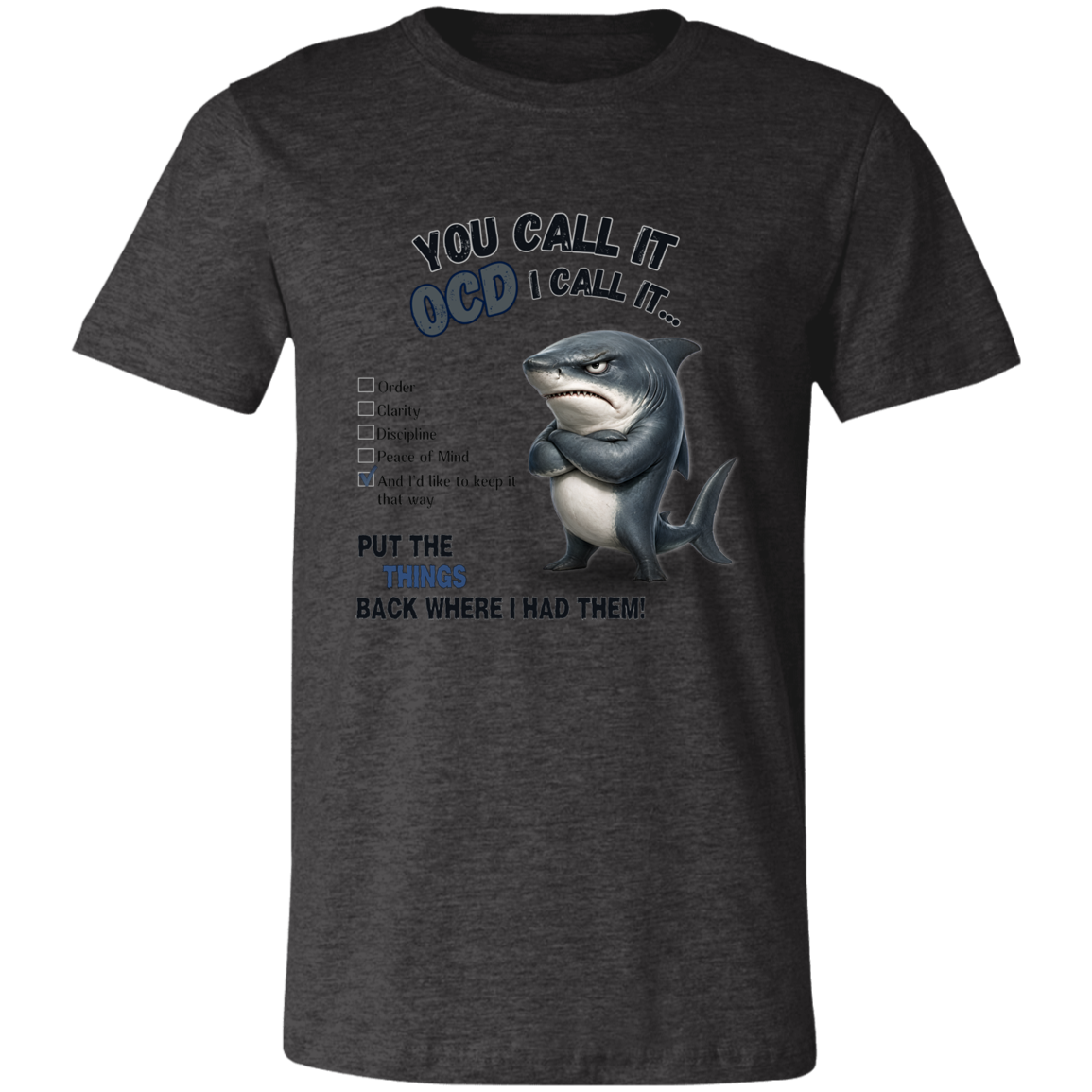 A unisex t-shirt in dark grey heather, featuring a graphic print of a shark with crossed arms and the text 'YOU CALL IT OCD I CALL IT.' along with a checklist and the phrase 'PUT THE TRICKS BACK WHERE I HAD THEM!' on a transparent 