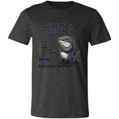 A unisex t-shirt in dark grey heather, featuring a graphic print of a shark with crossed arms and the text 'YOU CALL IT OCD I CALL IT.' along with a checklist and the phrase 'PUT THE TRICKS BACK WHERE I HAD THEM!' on a transparent 