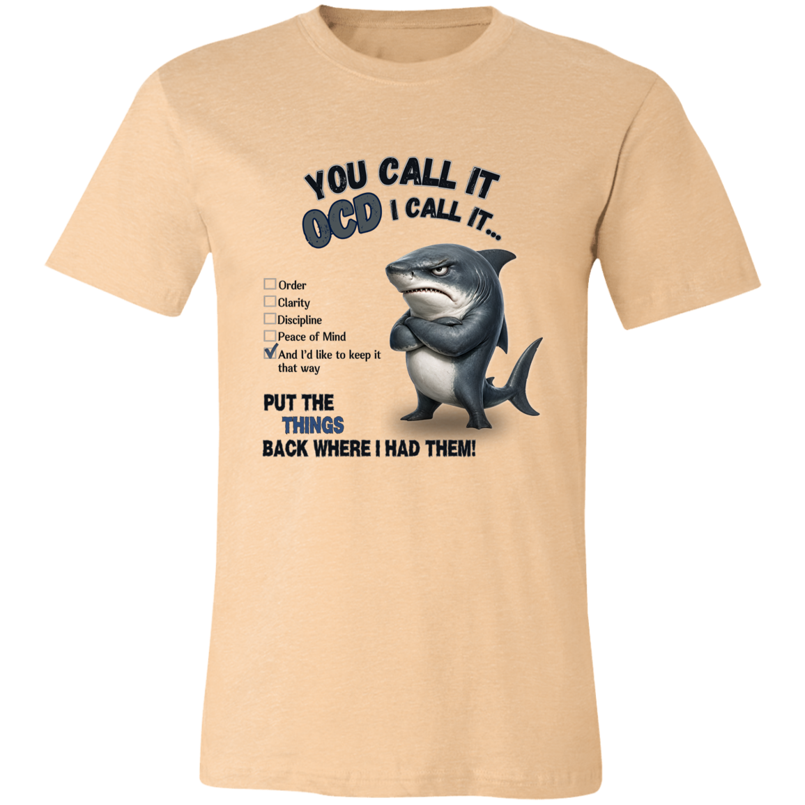 A unisex t-shirt in heather sand dune, featuring a graphic print of a shark with crossed arms and the text 'YOU CALL IT OCD I CALL IT.' along with a checklist and the phrase 'PUT THE TRICKS BACK WHERE I HAD THEM!' on a transparent 