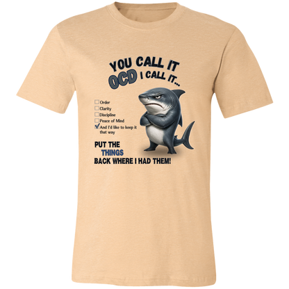 A unisex t-shirt in heather sand dune, featuring a graphic print of a shark with crossed arms and the text 'YOU CALL IT OCD I CALL IT.' along with a checklist and the phrase 'PUT THE TRICKS BACK WHERE I HAD THEM!' on a transparent 