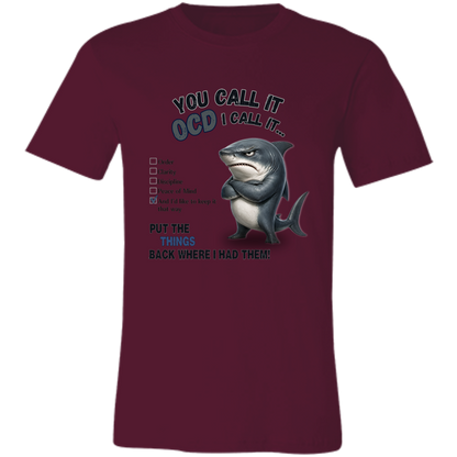 A unisex t-shirt in maroon, featuring a graphic print of a shark with crossed arms and the text 'YOU CALL IT OCD I CALL IT.' along with a checklist and the phrase 'PUT THE TRICKS BACK WHERE I HAD THEM!' on a transparent 