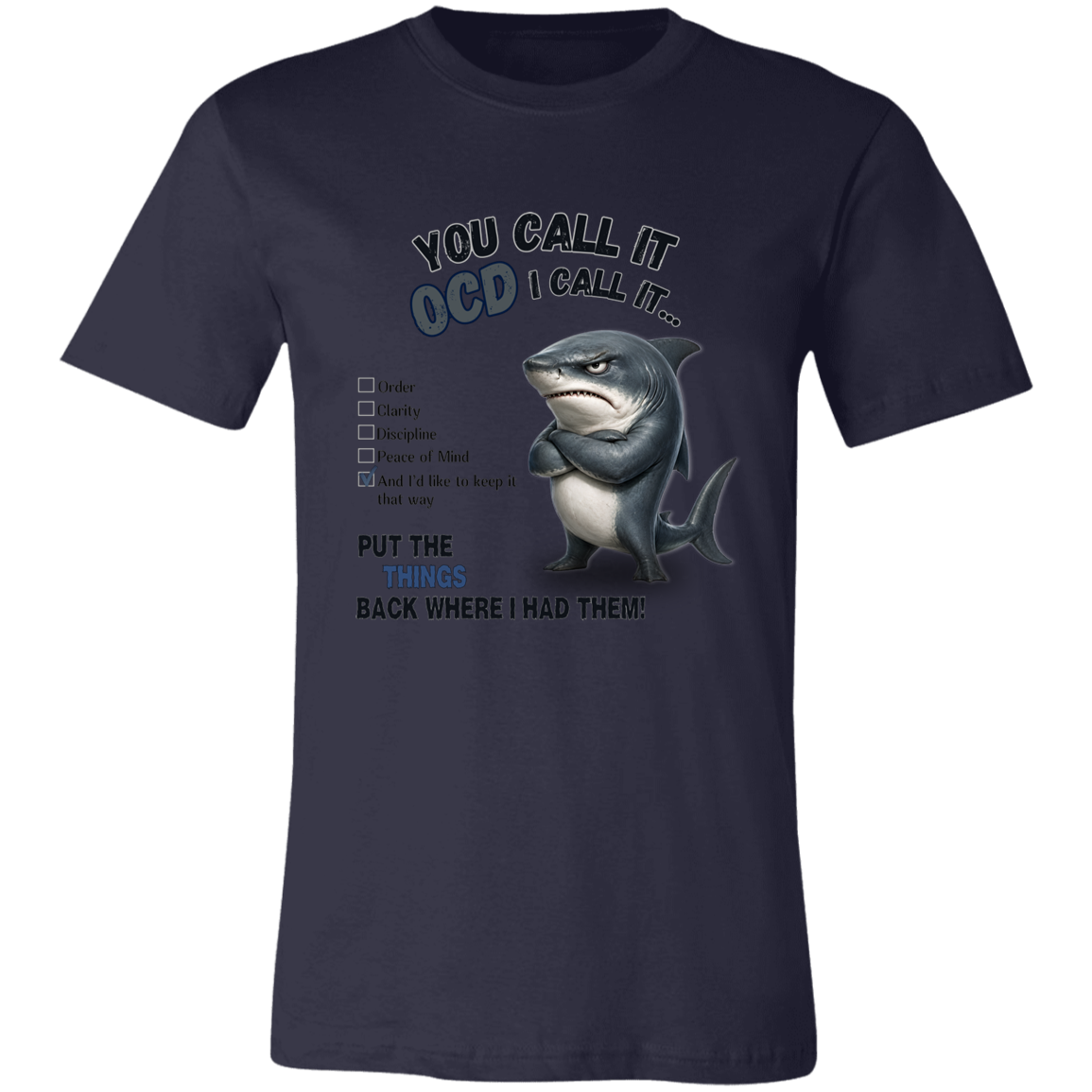 A unisex t-shirt in navy, featuring a graphic print of a shark with crossed arms and the text 'YOU CALL IT OCD I CALL IT.' along with a checklist and the phrase 'PUT THE TRICKS BACK WHERE I HAD THEM!' on a transparent 