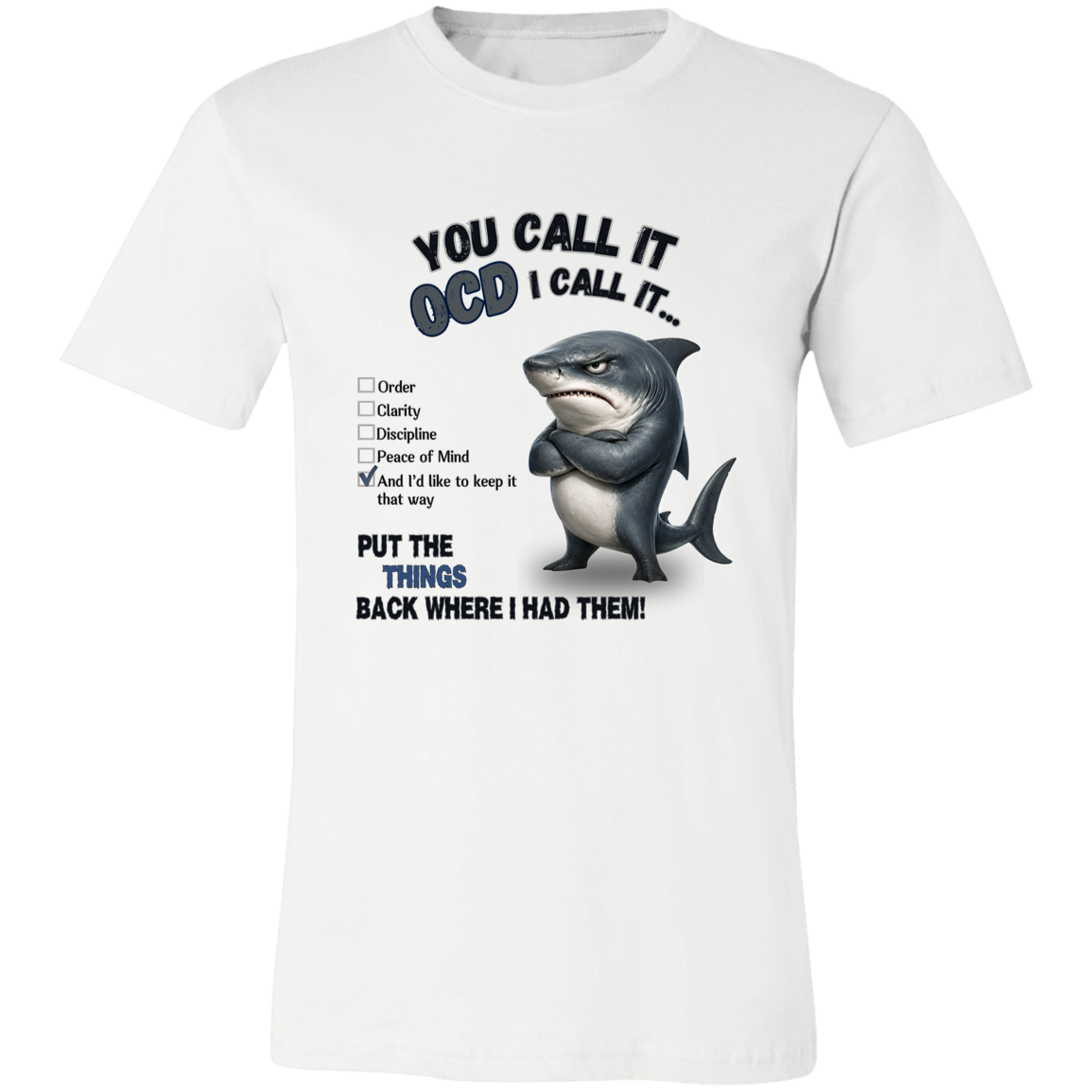 A unisex t-shirt in white, featuring a graphic print of a shark with crossed arms and the text 'YOU CALL IT OCD I CALL IT.' along with a checklist and the phrase 'PUT THE TRICKS BACK WHERE I HAD THEM!' on a transparent 
