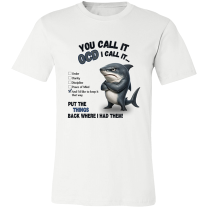 A unisex t-shirt in white, featuring a graphic print of a shark with crossed arms and the text 'YOU CALL IT OCD I CALL IT.' along with a checklist and the phrase 'PUT THE TRICKS BACK WHERE I HAD THEM!' on a transparent 