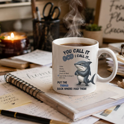 A white ceramic mug with a humorous OCD-themed shark graphic and text, shown on a desk with notebooks, a pen, a candle, and steam rising from the mug.