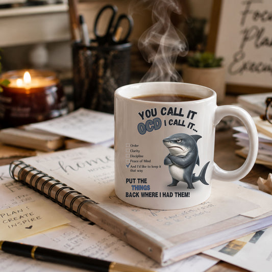 A white ceramic mug with a humorous OCD-themed shark graphic and text, shown on a desk with notebooks, a pen, a candle, and steam rising from the mug.